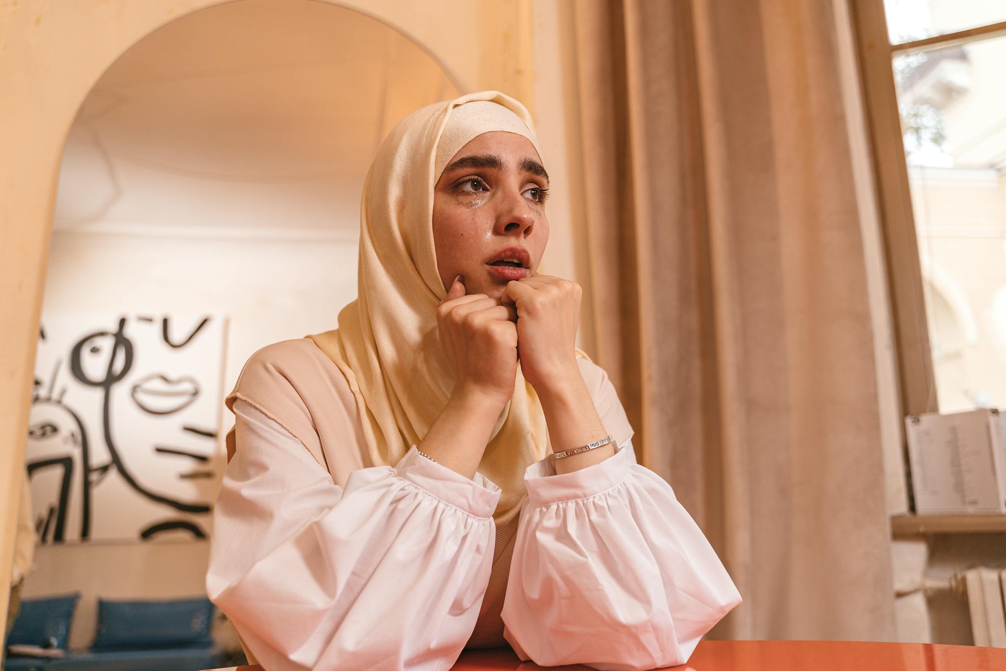 A woman in a hijab looks contemplative and emotional indoors, conveying a sense of introspection.